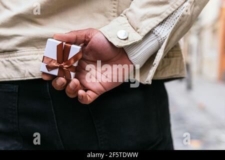 Rückansicht des anonymen Mannes mit der Hand hinter dem Rücken Halten kleine Geschenkbox mit Band in der Stadt Stockfoto