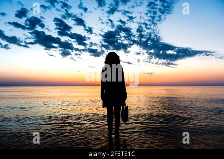 Rückansicht der Silhouette der nicht erkennbaren Frau im Wasser stehen Vom Meer und bewundern spektakuläre Aussicht auf den bunten Sonnenuntergang Himmel Im Sommer Stockfoto