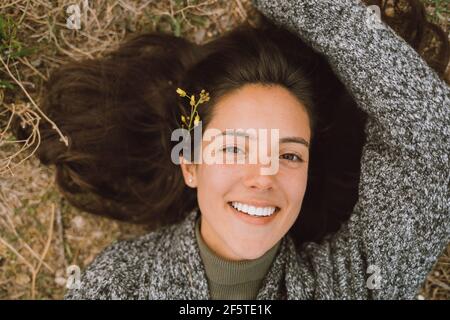 Draufsicht auf fröhliche junge Frau in warmer Strickmode Mit blühenden Blume im Haar Blick auf Kamera beim Genießen Frühlingstag während der Reise in der Natur Stockfoto