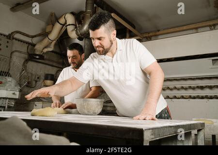 Von unten reifen bärtigen ethnischen Bäcker bilden Brot aus Teig Am Tisch mit Mehl und Schüssel in der Bäckerei Stockfoto