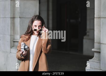 Stilvolle Frau in Mantel und Sonnenbrille stehen mit Vintage-Foto Kamera auf der Straße und Blick auf die Kamera Stockfoto