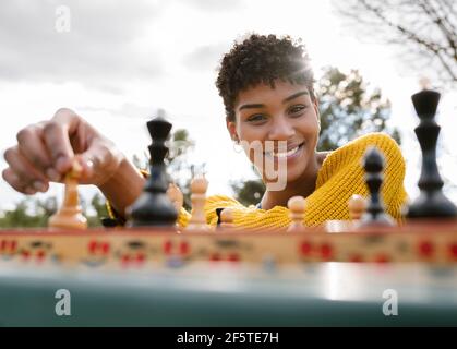 Von unten der optimistischen afroamerikanischen Frau, die am Tisch sitzt Im Park und Schach spielen, während Sie die Kamera betrachten Stockfoto