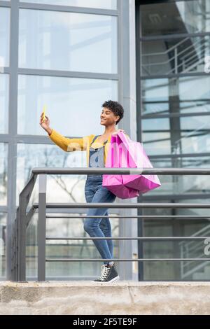 Inhalt Afrikanisch-amerikanische Frau, die in der Nähe des Glasgebäudes mit Einkaufsmöglichkeiten steht Taschen und Selbstschuss auf dem Smartphone Stockfoto