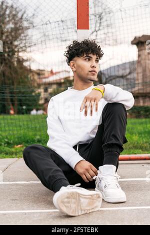 Junger afroamerikanischer Mann in trendigen Kleidern sitzt auf Basketball Spielplatz und Blick weg Stockfoto