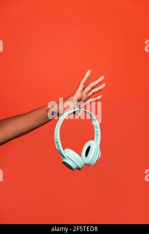 Crop anonyme afroamerikanische Frau zeigt drahtlose Kopfhörer auf ausgestreckt Hand vor rotem Hintergrund im Studio Stockfoto