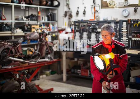 Qualifizierte ältere männliche Mechaniker setzen glänzende elektrische Lampe in der Nähe des Arbeitsplatzes Während der Vorbereitung für die Reparatur in der Garage mit Motorrad und Instrumente Stockfoto