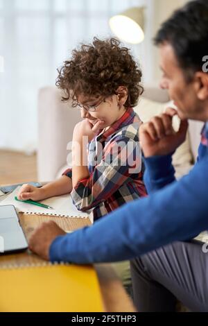 Zeit zum Lernen. Neugieriger kleiner lateinischer Schuljunge, der zusammen mit seinem Vater Hausaufgaben macht, auf Papier schreibt, während er zu Hause am Schreibtisch sitzt Stockfoto