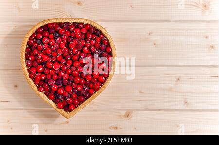 Ein Weidengeschirr in Herzform, gefüllt mit gefrorenen roten Preiselbeeren. Auf der hellen Holzplatte links. Stockfoto