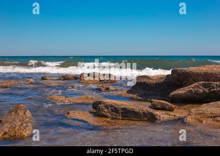 Meeresküste mit einer blauen Welle, die gegen die Steine am Strand schlägt. Meer sonnige Sommerlandschaft. Stockfoto