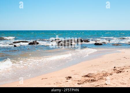 Meeresküste mit blauem Wasser gegen Steine und einem Sandstrand. Meer sonnige Sommerlandschaft. Stockfoto
