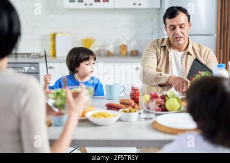 Vater mittleren Alters im Gespräch mit seinem Kind beim Frühstück am Morgen. Lateinische Familie genießen zusammen Essen, sitzen am Tisch in der Küche Stockfoto