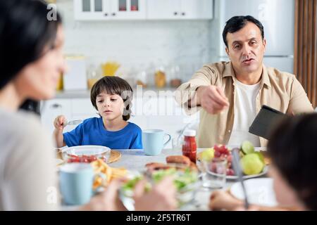 Vater mittleren Alters chattet mit Kindern, mit Tablet-pc beim Essen in Innenräumen. Lateinische Familie genießen zusammen Essen, sitzen am Tisch in der Küche Stockfoto