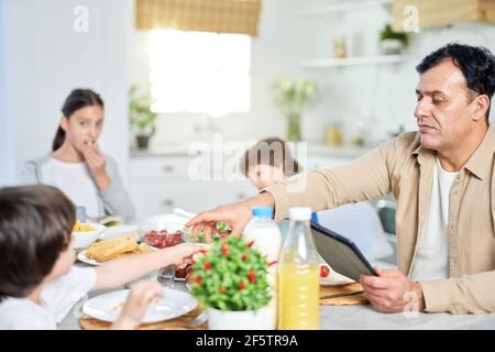 Vater mittleren Alters genießen zusammen mit seiner Familie Essen, mit digitalen Tablet, während am Tisch in der Küche zu Hause sitzen Stockfoto