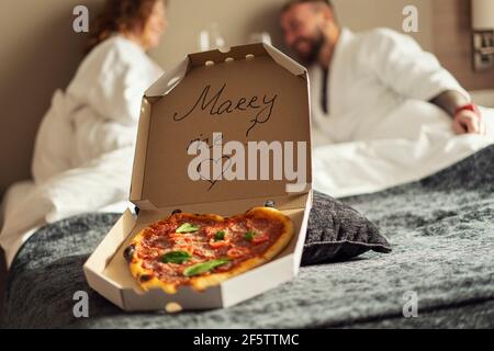 Marry Me, Schriftzug auf Pizzaschachtel auf verschwommenem Hintergrund verliebte sich in Gläser Wein und weiße Bademäntel Stockfoto