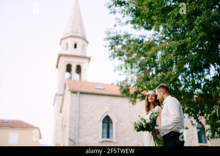 Braut und Bräutigam gehen durch die Altstadt von Budva, der Bräutigam umarmt die Braut sanft und hält einen Strauß in der Hand Stockfoto