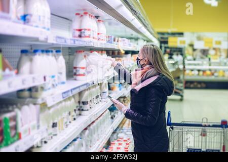 Blonde Frau in Schutzmaske Milchprodukte aus dem Regal im Supermarkt nehmen. Stockfoto