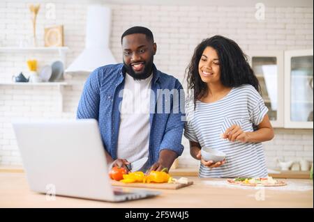 Portrait von jungen glücklichen lateinischen Paar mit Laptop beim Kochen in der Küche zu Hause, Blick auf den Bildschirm, beobachten kulinarischen Tutorial Lektion, bereitet ihr Lieblingsrezept der gesunden Gemüse Pizza Stockfoto