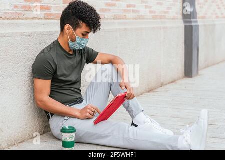 Porträt eines attraktiven lateinamerikanischen Mannes, der mit einer roten Tafel auf dem Boden der Straße sitzt. Er kleidet sich lässig und trägt eine schützende Gesichtsmaske. Er hat eine wiederverwendbare Kaffeetasse in seiner Nähe Stockfoto