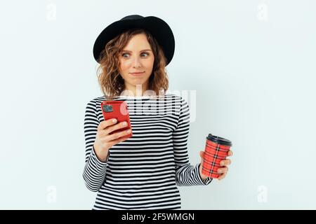 Schockiert verwirrt junge Frau trägt schwarzen Hut und gestreiftes Hemd halten rote Tasse Kaffee Blick in ihr Smartphone, weißen Studio Hintergrund. Stockfoto