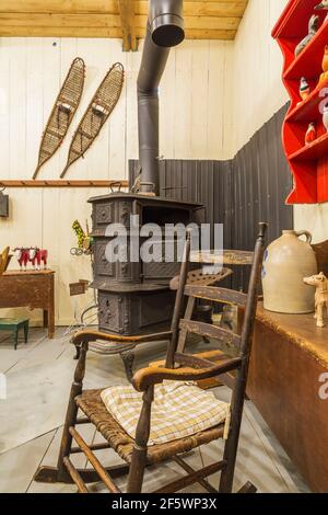 Antiker hölzerner Schaukelstuhl, alter schwarzer gusseiserner Holzofen und Gegenstände, die in einer alten Holzplankenscheune ausgestellt sind Stockfoto