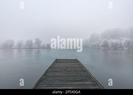 Gefrorener See im Winter, mit hölzernen Pier, und Bäume von Schnee bedeckt, schönen nebligen Sonnenuntergang auf dem Wald. Schöne saisonale Winterlandschaft friedlich Stockfoto