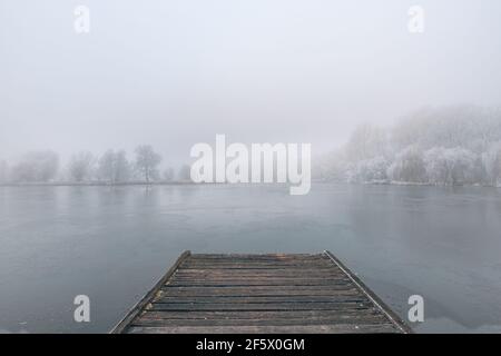 Gefrorener See im Winter, mit hölzernen Pier, und Bäume von Schnee bedeckt, schönen nebligen Sonnenuntergang auf dem Wald. Schöne saisonale Winterlandschaft friedlich Stockfoto