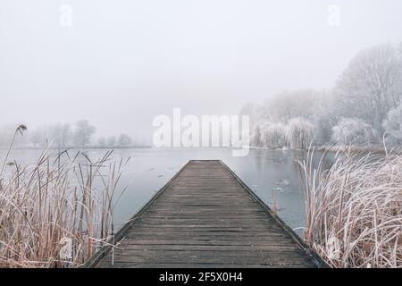 Gefrorener See im Winter, mit hölzernen Pier, und Bäume von Schnee bedeckt, schönen nebligen Sonnenuntergang auf dem Wald. Schöne saisonale Winterlandschaft friedlich Stockfoto