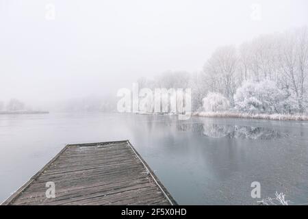 Gefrorener See im Winter, mit hölzernen Pier, und Bäume von Schnee bedeckt, schönen nebligen Sonnenuntergang auf dem Wald. Schöne saisonale Winterlandschaft friedlich Stockfoto