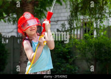 Kleines Feuerwehrmädchen, kleines Kind in einem roten Spielzeug Feuerwehrhelm mit einer Wasserpistole Wasser in die Luft sprühen, Außenportrait, zukünftige Job Träume, Stockfoto