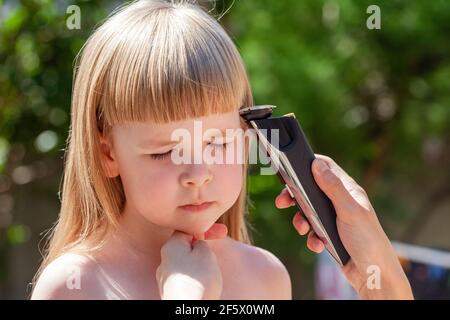 Mutter, Elternteil, der mit einem elektrischen Haarschneider die Haare ihrer kleinen Tochter schneidet, Porträt im Freien. Elektrische Haarschneider Amateur Haarschnitt bei Stockfoto