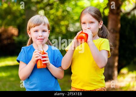 Zwei glückliche junge fröhliche kleine Mädchen im Schulalter halten geöffnete Glasflaschen voll von gesunden Orangensaft Obst trinken lächelnd, im Freien portra Stockfoto