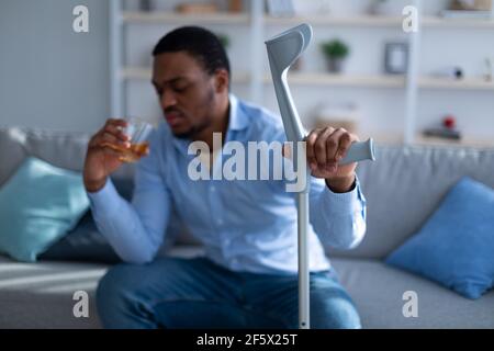 Schwarzer Mann mit Krücke Gefühl deprimiert aufgrund seiner Verletzung, Alkohol zu trinken, um seine Schmerzen zu Hause zu lindern, selektive Konzentration Stockfoto