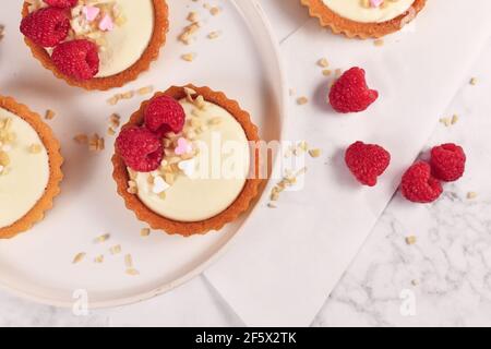 Blick von oben auf kleine Tartlet-Gebäck mit weißer Sahne, gekrönt mit Himbeerfrüchten und Mandel- und herzförmigen Zuckerstreuseln Stockfoto