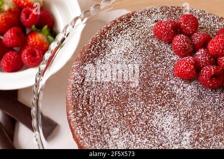 Hausgemachte Schokoladenkuchen serviert auf einem eleganten Glasteller mit Himbeeren oben und eine Schüssel Erdbeeren daneben IT Stockfoto