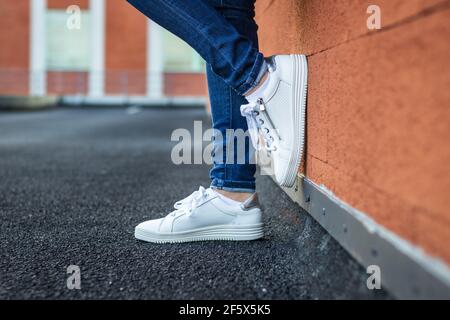 Frau mit Leder weiß Sneaker lehnte Wand in der Stadt, Nahaufnahme Seitenansicht zu weiblichen Beinen tragen Jeans und Sportschuh. Modekonzept Stockfoto