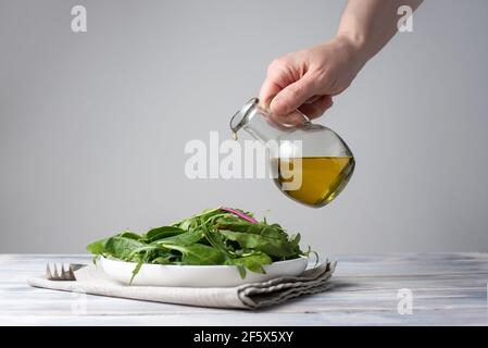 Weibliche Hand Gießen Olivenöl in einen Teller mit einem Salat von Blattgemüse auf weißem Holzhintergrund, vegetarisches Essen, gesunde Ernährung, Nahaufnahme Stockfoto