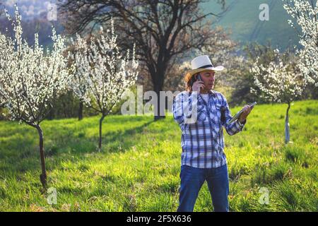 Landwirt mit Smartphone und digitale Tablette in blühenden Obstgarten. Landwirtschaft und moderne Technik Konzept. Agronom im Gespräch mit dem Handy Stockfoto