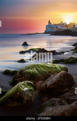 Schöner und farbenfroher Sonnenuntergang am Strand mit Moschus auf Felsen und glattem Wasser in Sitges, Katalonien, Spanien Stockfoto