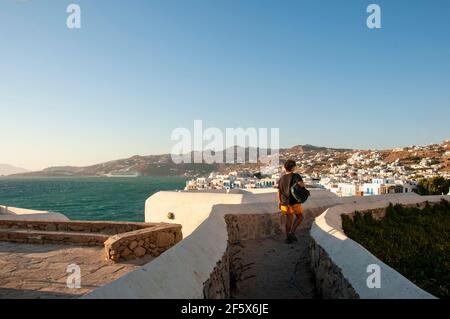 Ein junger Mann mit einem Rucksack auf der Schulter geht entlang einer malerischen Straße auf der Insel Mykonos in Griechenland. Im Hintergrund das Meer und das Dorf mit Stockfoto