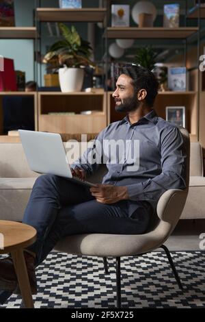 Indischer Geschäftsmann schaut weg, sitzt auf einem Stuhl im modernen Büro mit Laptop. Stockfoto
