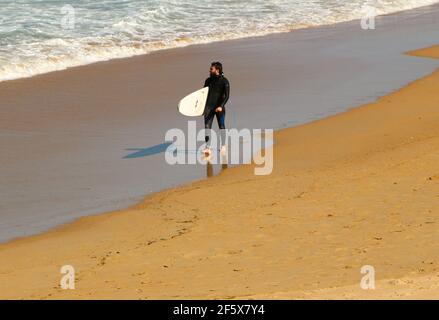 Ein eingeflügeliger Surfer, der an einem frischen, windigen Frühlingsnachmittag in Sardinero mit einem weißen Surfbrett am Strand entlang geht Santander Cantabria Spanien Stockfoto