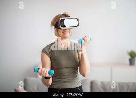 Sportliche reife Frau in VR-Headset arbeiten ihre Bizeps Mit Kurzhanteln während des inländischen Trainings Stockfoto
