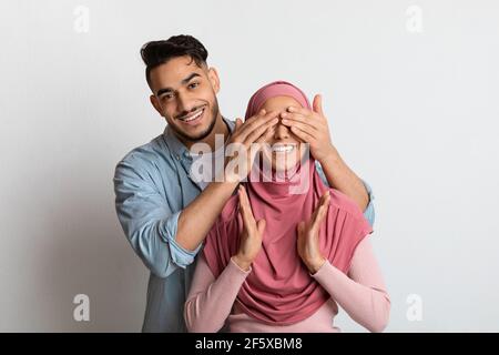 Liebevoller arabischer Mann, der seine aufgeregte Frau in Hijab überraschte und ihre Augen verhüllte Stockfoto