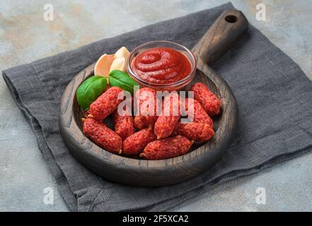 Holzplatte mit Knoblauch-Mini-Würstchen, Knoblauch, Basilikum und Sauce auf grauem Hintergrund. Wurstsnacks. Seitenansicht, Nahaufnahme. Stockfoto