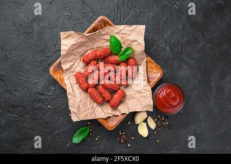 Ein Holzteller mit kleinen geräucherten Würstchen, Knoblauch und Basilikum auf dunklem Grund. Das Konzept der Wurstwaren. Stockfoto