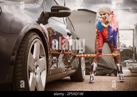 Moskau, Russland - 06. Juli 2020: Harley Quinn trifft mit einem Baseballschläger auf ein geparktes schwarzes Auto. Es ist eine Delle im Auto, das Auto ist beschädigt. Cosplay, Mädchen verkleidet als Charakter Kostüm Stockfoto