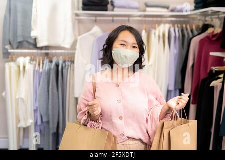 Porträt von glücklichen asiatischen jungen Frau in Schutzmaske halten Käufe und Blick auf die Kamera im Bekleidungsgeschäft stehen Stockfoto