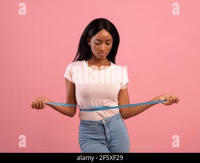 Gewichtsverlust. Traurige schwarze Frau, die ihre Taille mit Klebeband misst, unzufrieden mit den Körperparametern auf pinkem Studiohintergrund Stockfoto