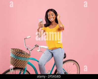 Hübsche schwarze Frau auf Vintage-Fahrrad unter Selfie mit Smartphone Über rosa Studiohintergrund Stockfoto