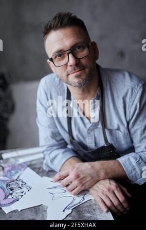 Bärtiger männlicher Künstler mittleren Alters in einer Brille, die auf einem Hintergrund des Arbeitsplatzes sitzt. Linolschnitt Meister in Schürze lächelnd und gerade in der Kamera und lachend. Hochwertige Fotos Stockfoto